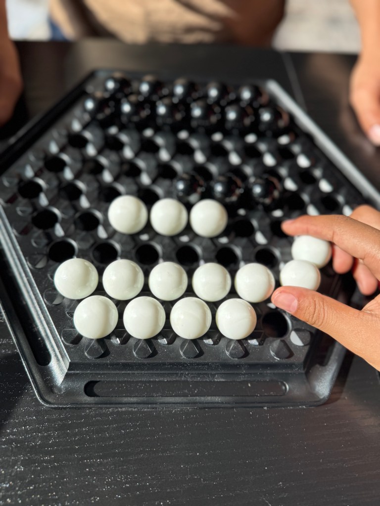 A close-up view of a hexagonal board game featuring black and white marbles, with a hand about to move a white marble.