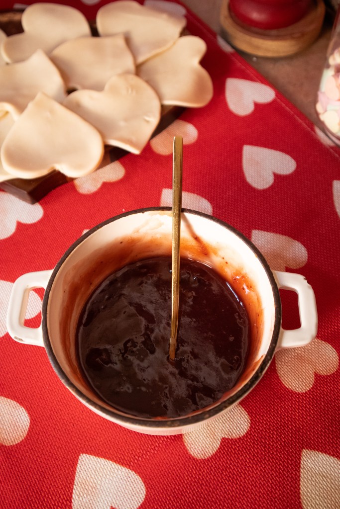 A small pot of strawberry sauce with a spoon, resting on a red tablecloth decorated with white hearts, beside heart-shaped pie crust.