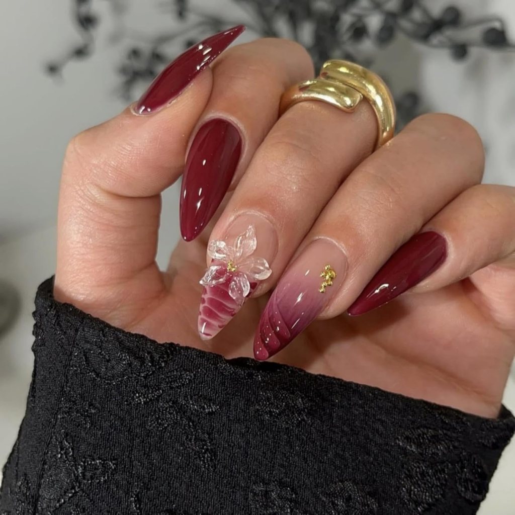 Close-up of a hand with long, pointed burgundy nails featuring a decorative crystal flower and a gradient effect on one nail, all highlighted against a dark clothing background.