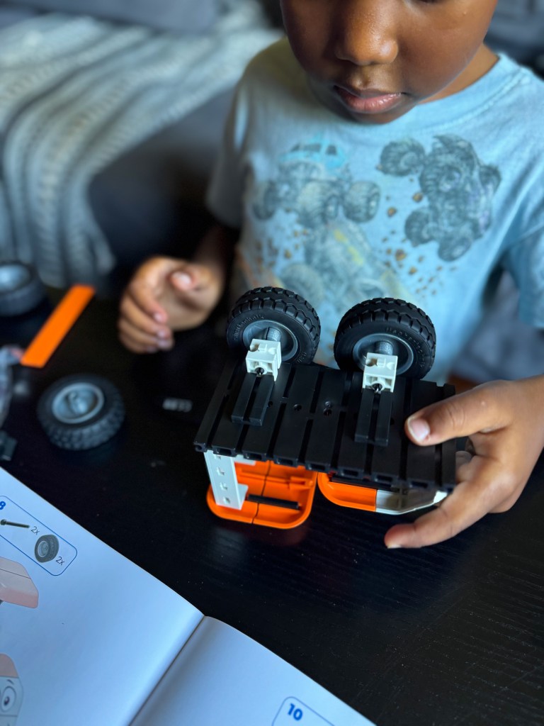 A child assembling a toy vehicle using building blocks, with a guidebook and scattered toy parts visible on a black table.