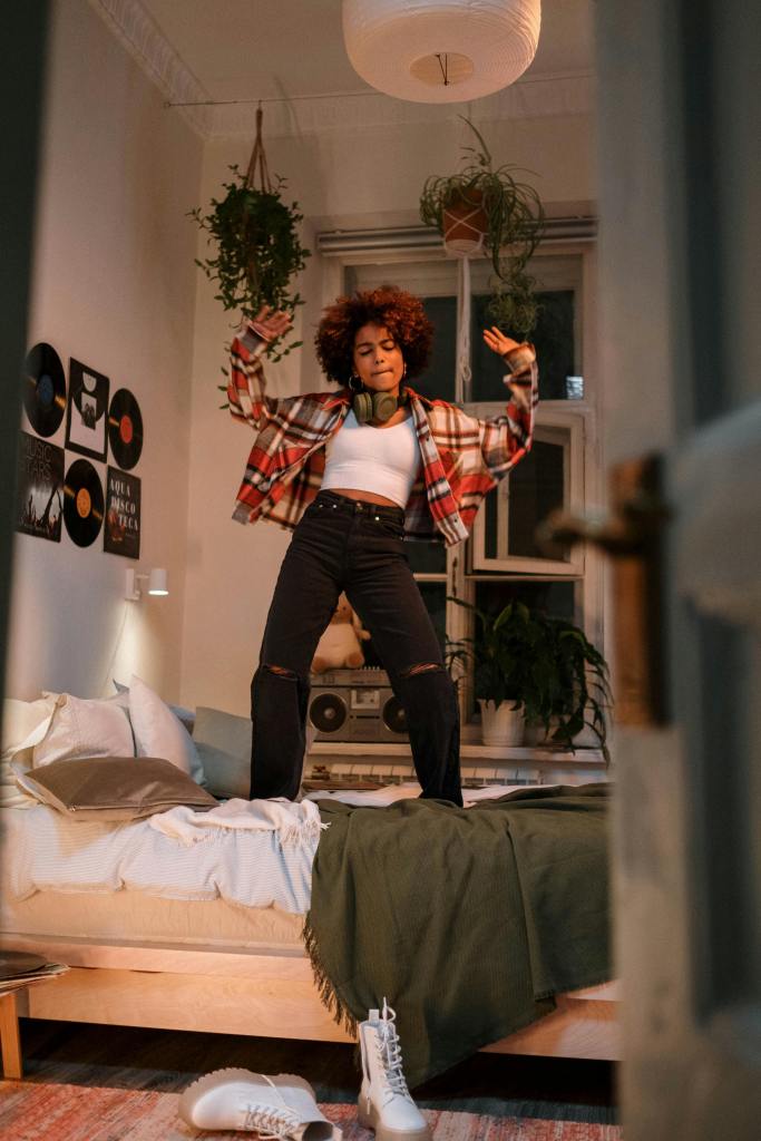 A person with curly hair dances joyfully on a bed in a cozy, warmly lit room decorated with plants and records.