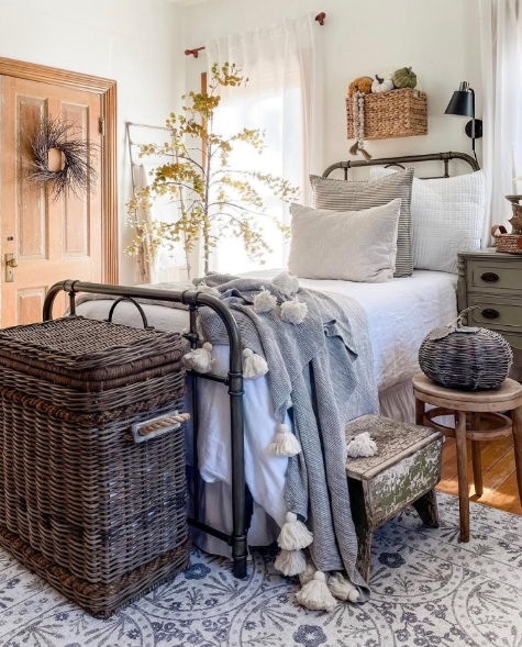A cozy bedroom featuring a metal bed frame with soft bedding, decorative pillows, and a warm gray blanket. A rustic wicker basket and stool accompany the bed, while a small tree adds a touch of natural beauty. The room is bright and inviting, showcasing warm tones and textured decor.