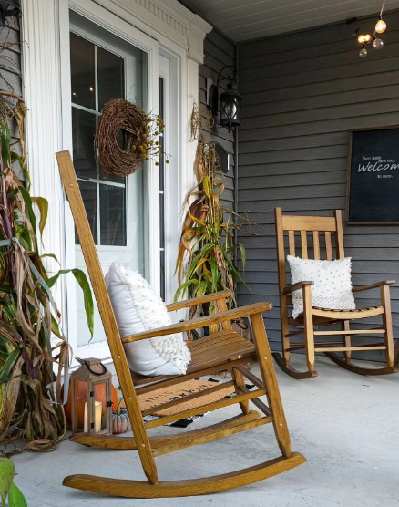 A cozy porch featuring two wooden rocking chairs, one adorned with a white pillow, alongside a decorative wreath and corn stalks. Lanterns and a welcome sign enhance the inviting autumn atmosphere.