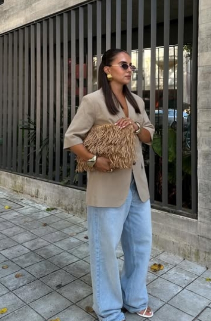Woman in oversized beige blazer, wide-leg light wash jeans, and straw textured clutch for a chic fall outfit.