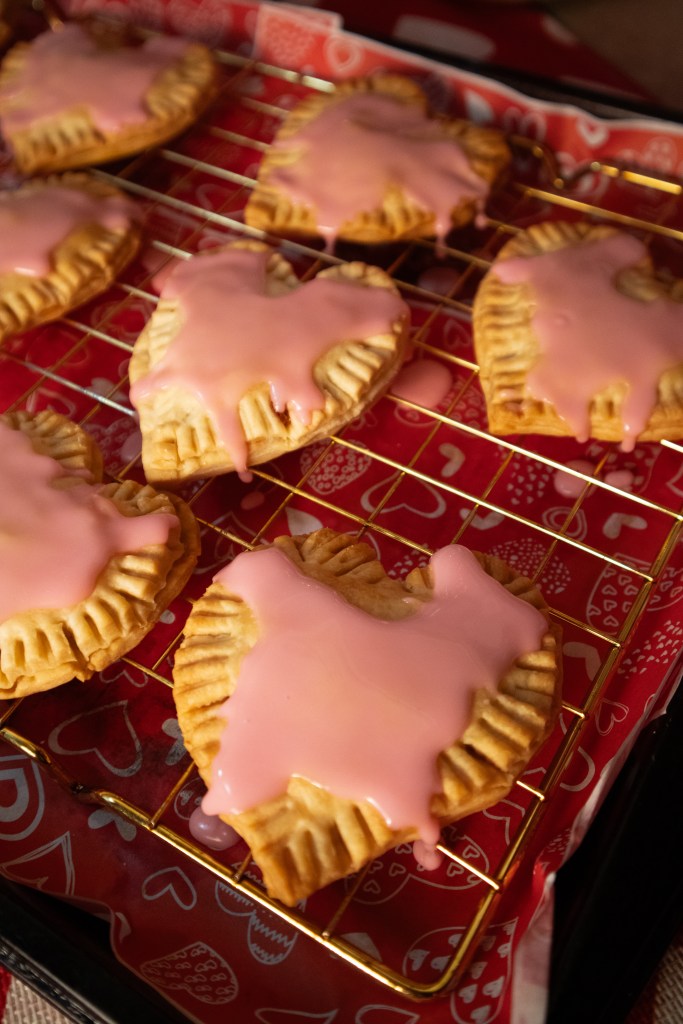 Freshly baked heart-shaped pop tarts with pink icing, displayed on a cooling rack lined with heart-patterned paper.