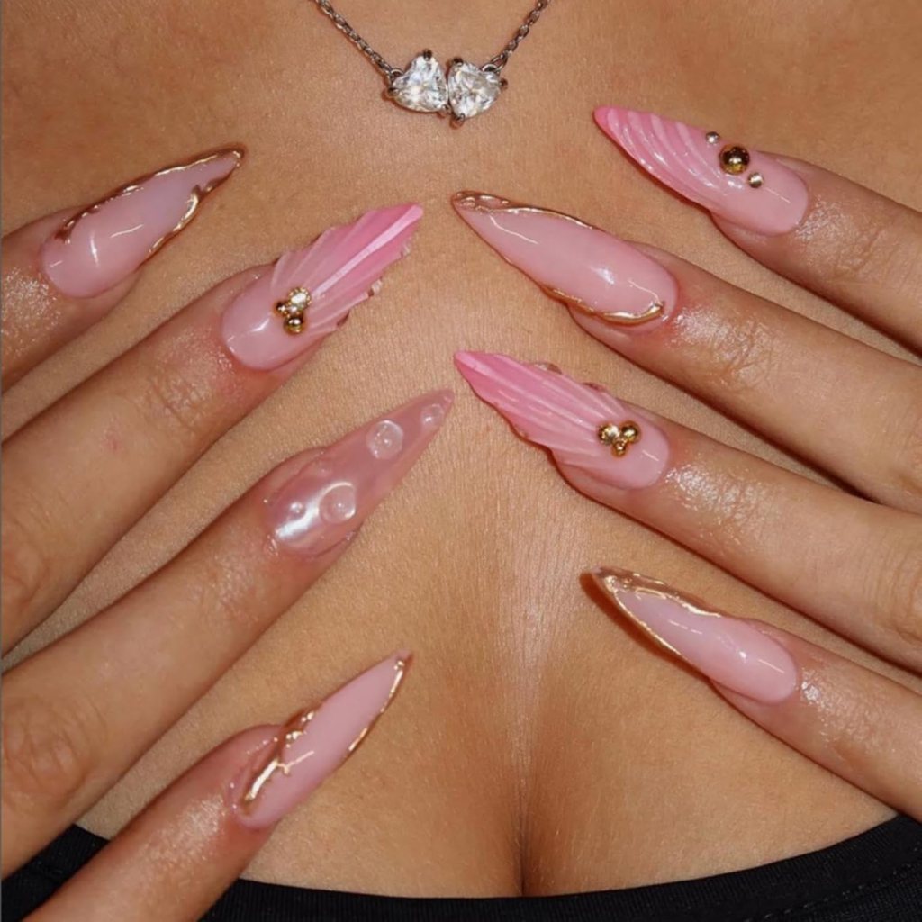 Close-up of elegantly manicured nails featuring a combination of pink and clear designs, adorned with gold accents and decorations, set against a neutral background.