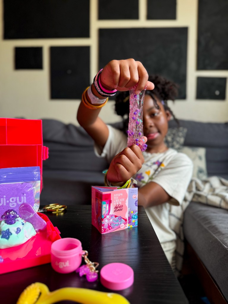A child holding a colorful, sticky slime in one hand while showcasing a pink box labeled 'Steps to Create Your Slime Scents' on a table with various crafting supplies.