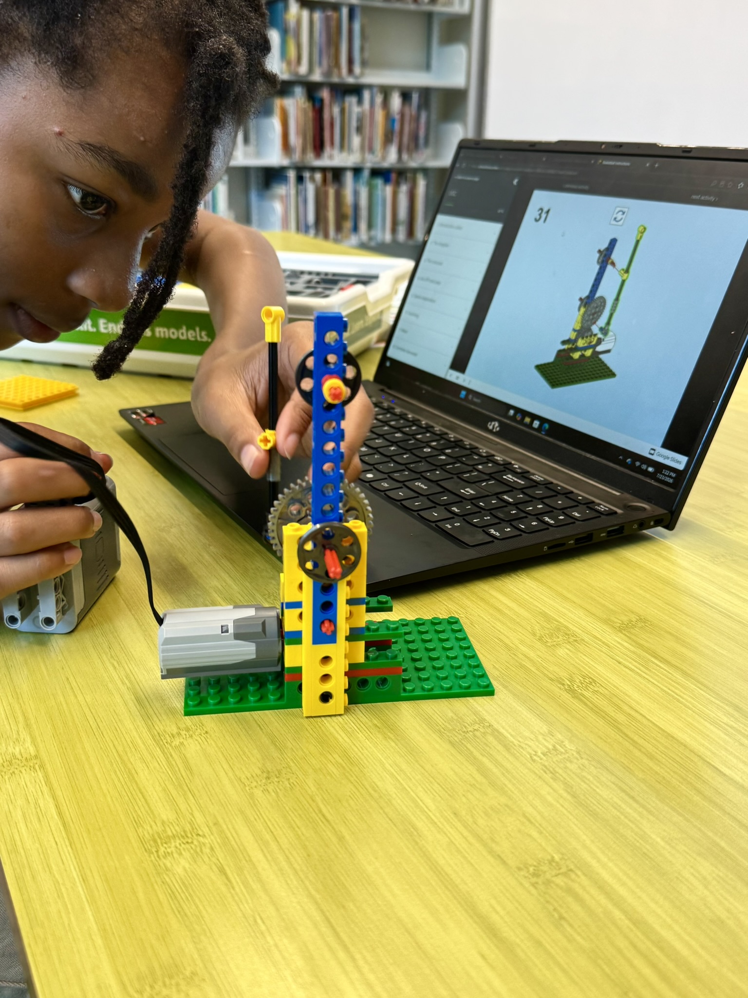Student assembling a LEGO robotic model using step-by-step instructions displayed on a laptop screen