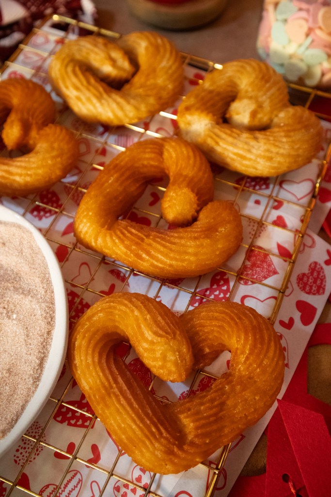 A golden brown batch of heart-shaped churros on a cooling rack, with a bowl of sugar nearby and a festive heart-patterned background.