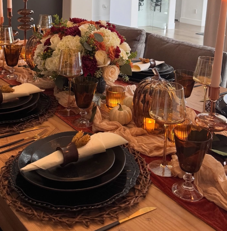 A beautifully styled Thanksgiving tablescape featuring dark plates, amber glassware, decorative pumpkins, and a floral centerpiece with rich autumn hues.
