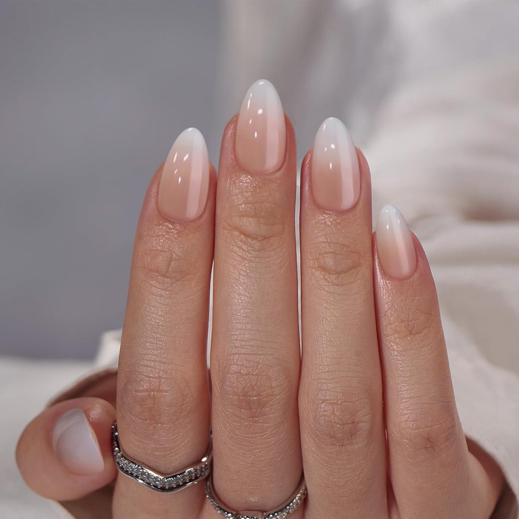 Close-up of a hand with elegantly manicured long nails featuring a gradient ombre effect, transitioning from soft pink at the base to white at the tips. The hand is adorned with multiple rings.