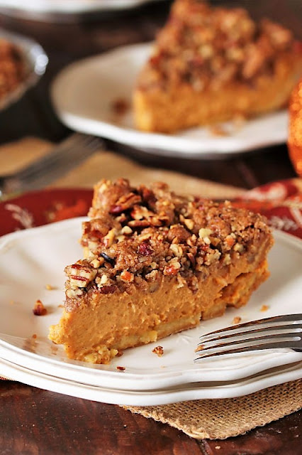 A slice of pumpkin pie with a pecan streusel topping served on a white plate, with a fork beside it, and autumn-themed decorations in the background.