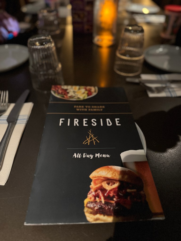 A close-up view of the Fireside restaurant menu at Great Wolf Lodge, featuring a burger image and the text 'All Day Menu'. The setting includes a dark wooden table with cutlery, glasses, and a candle in the background.