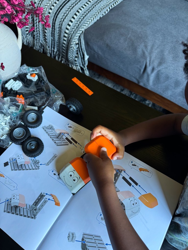 A child assembling a building toy, focusing on putting together two orange pieces from an instruction booklet, with wheels and other parts on the table.