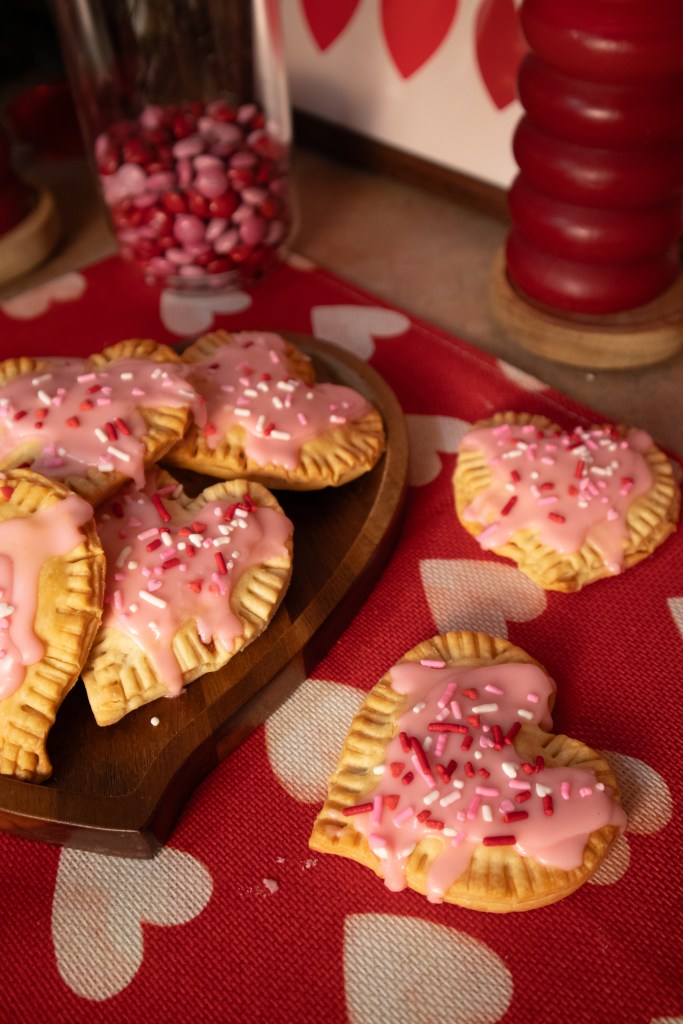 A wooden platter featuring heart-shaped pop tarts with pink icing and colorful sprinkles, set on a red tablecloth with heart patterns. A jar of red and pink candies is visible in the background.
