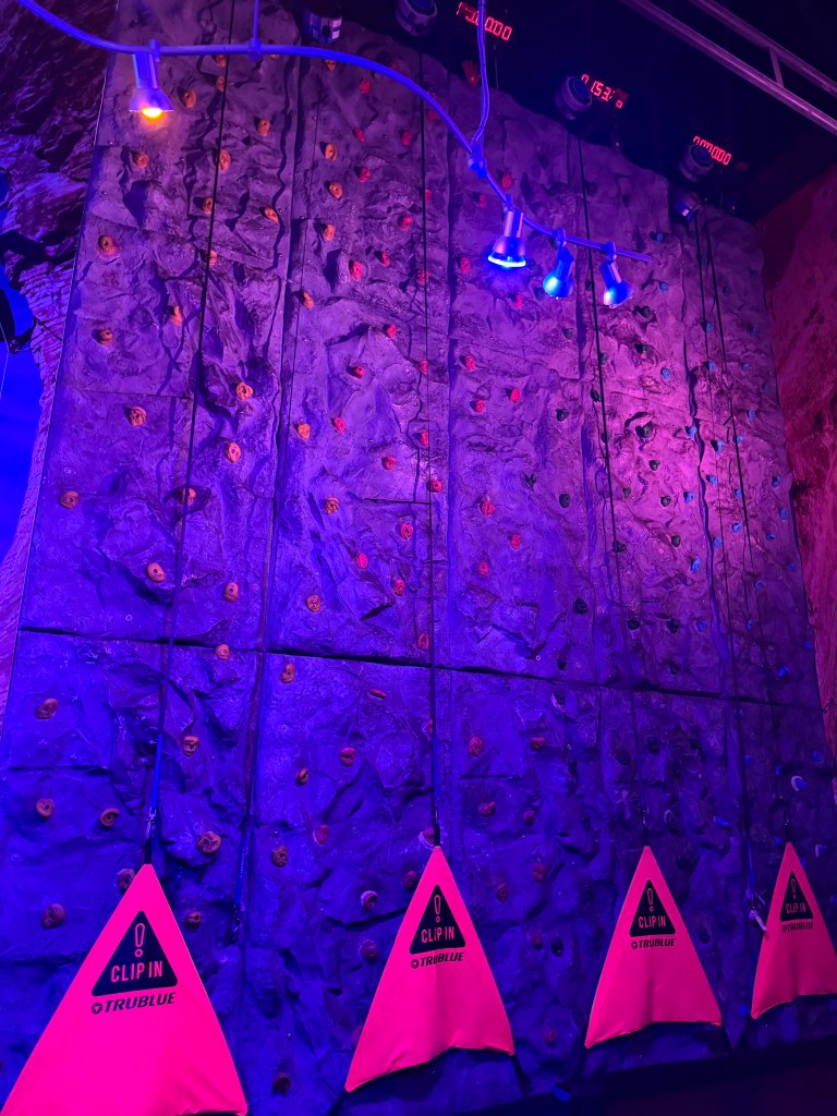Indoor climbing wall with colorful holds and illuminated with purple lights, featuring safety bags labeled 'Clip In TRUBLUE' at the base.