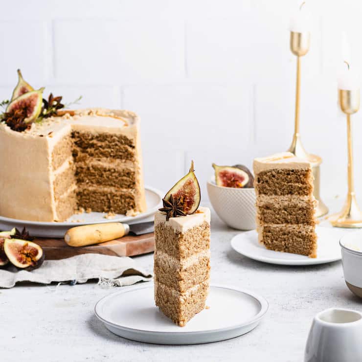 A slice of spiced layer cake topped with figs and star anise, served on a white plate, with the full cake in the background.