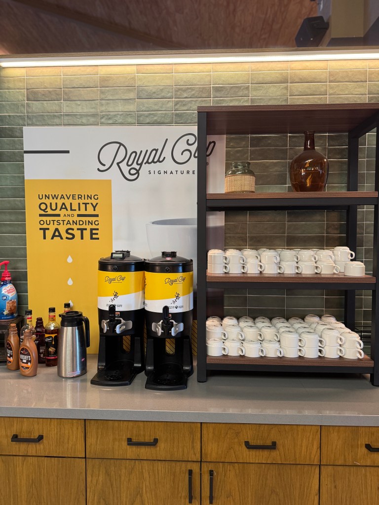 Coffee station featuring Royal Cup coffee dispensers, flavored syrups, and stacked cups on an organized countertop.