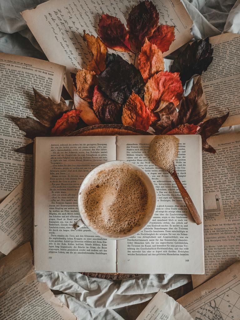A cozy autumn scene featuring a steaming cup of coffee on an open book, surrounded by colorful dried leaves and scattered pages.