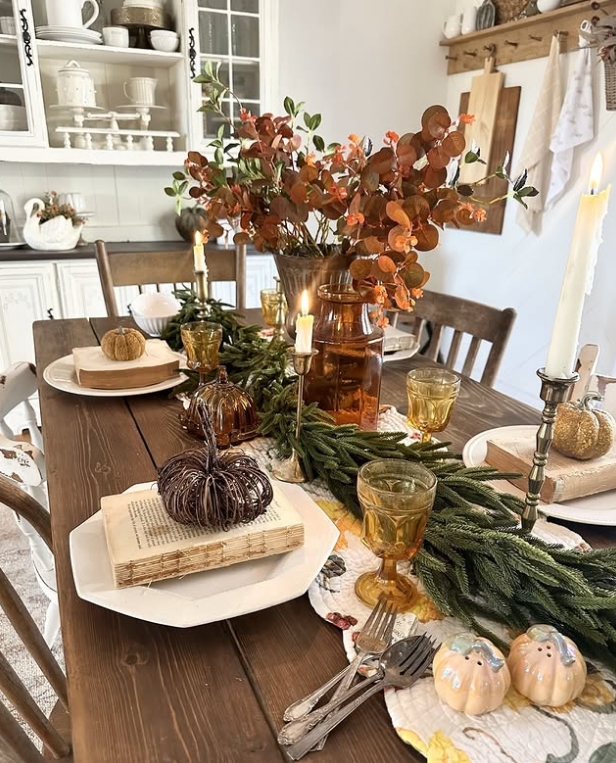 A beautifully styled Thanksgiving tablescape featuring warm autumn hues, decorative pumpkins, and layered natural elements. The table is adorned with golden glassware, flickering candles, and seasonal greenery, creating an inviting and cozy atmosphere.