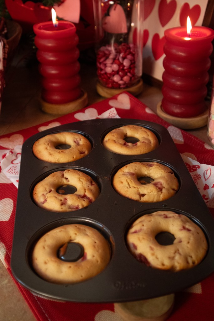 A baking tray with six baked doughnut-shaped treats, set against a festive backdrop featuring red candles and decorative hearts.