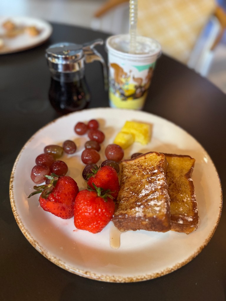 A plate of French toast with syrup, garnished with grapes, strawberries, and a small piece of cake, accompanied by a colorful beverage.
