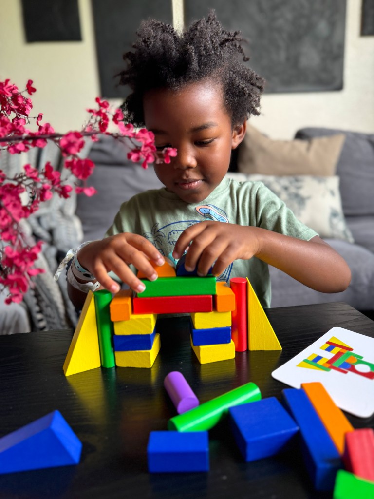A young child with curly hair is building a colorful structure using geometric blocks on a table. A branch with pink flowers is in the foreground, and a card with a similar design is visible beside the blocks.