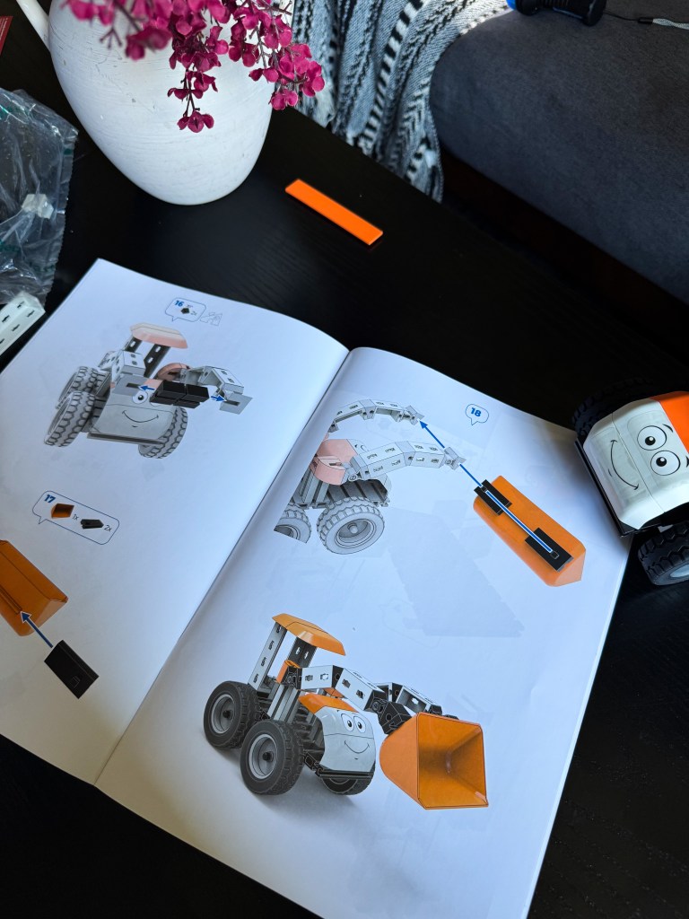 An open instruction manual showing steps to build a toy construction vehicle, with an image of the completed model on one page and scattered building blocks and tools on a black table.