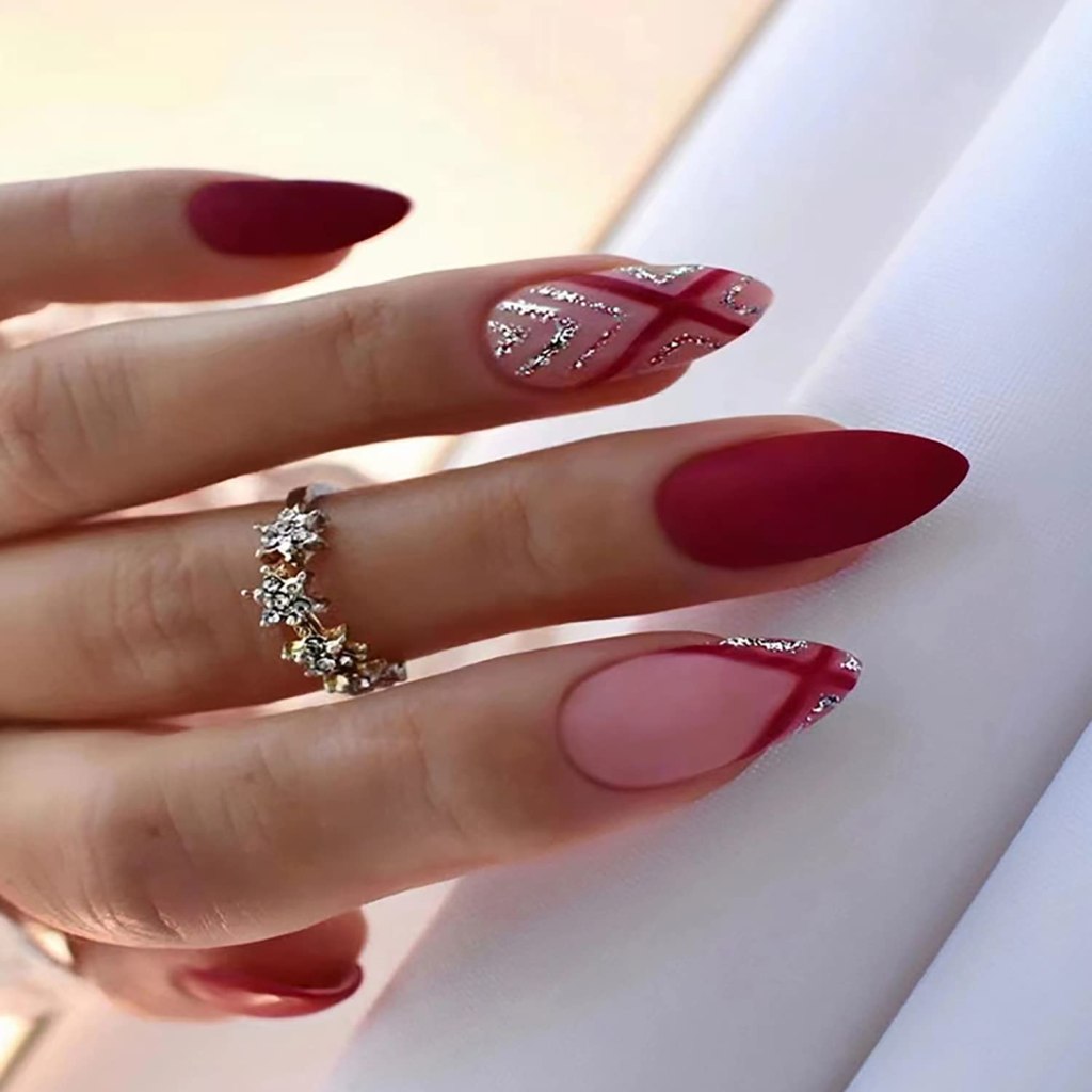 Close-up of a hand with manicured nails featuring a mix of deep red and pink polish. One nail has a glossy design with silver accents, while others are matte. A silver ring with floral designs is worn on the finger.