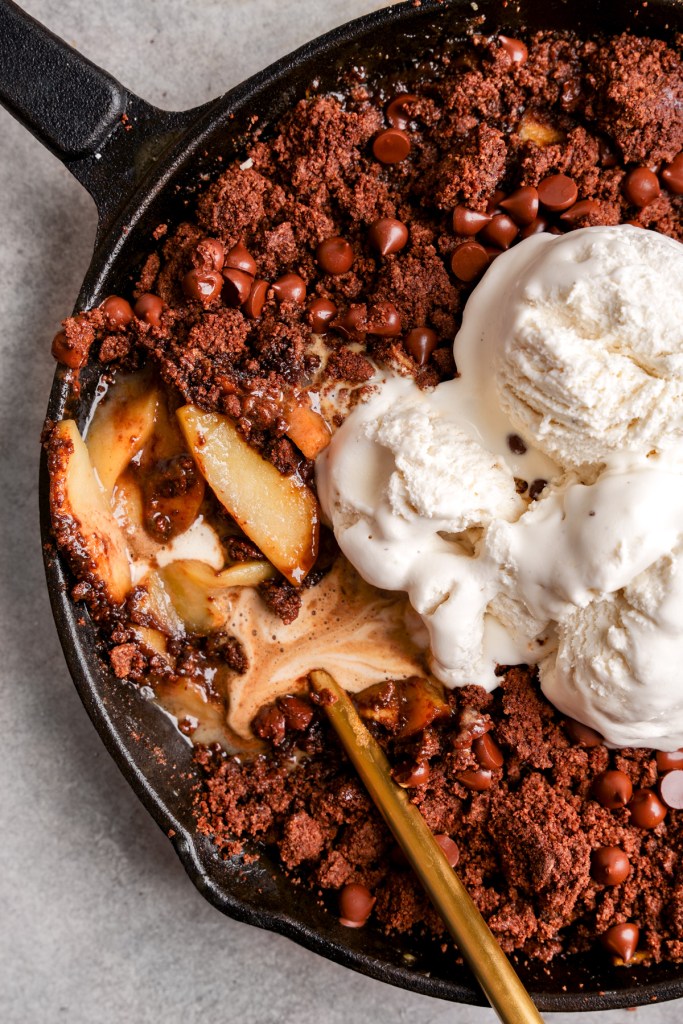 A close-up of a skillet dessert featuring baked apples topped with a chocolate crumble and vanilla ice cream.