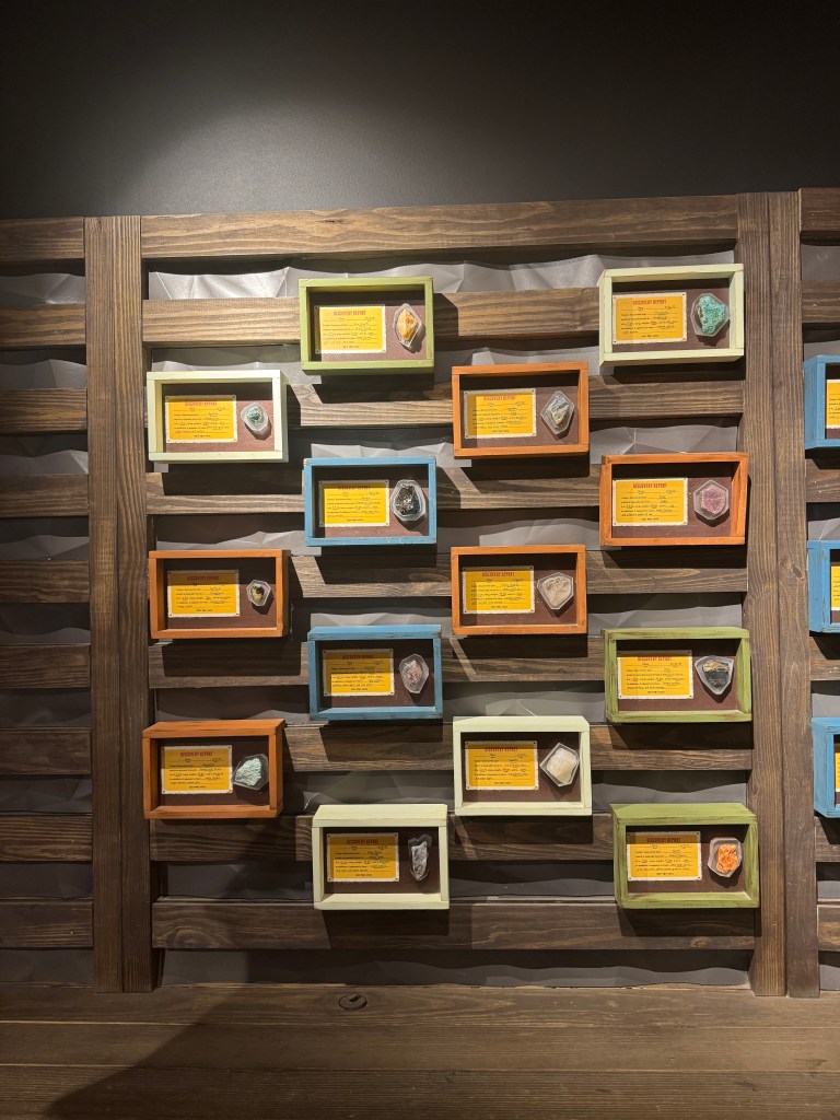 Display of various gemstones mounted in colorful frames on a wooden wall, with descriptions for each stone.
