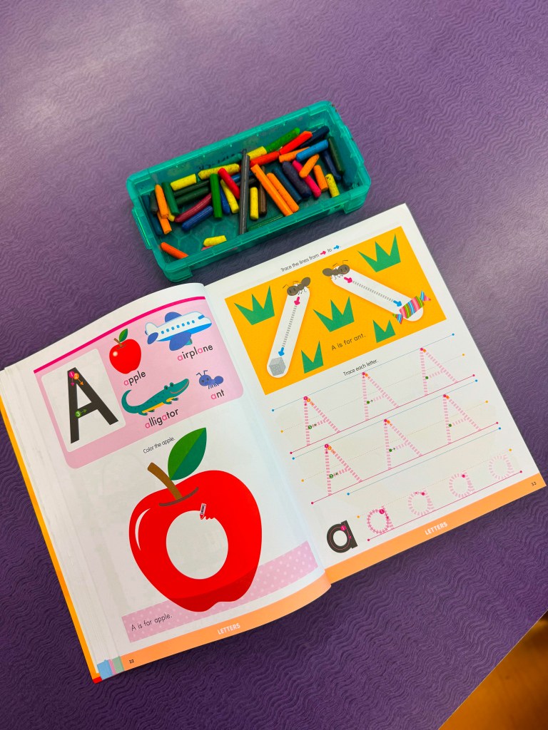 An open educational book page featuring letters and corresponding images, including an apple, airplane, alligator, and ant, along with coloring and tracing activities. A container with colorful crayons or markers is also visible.