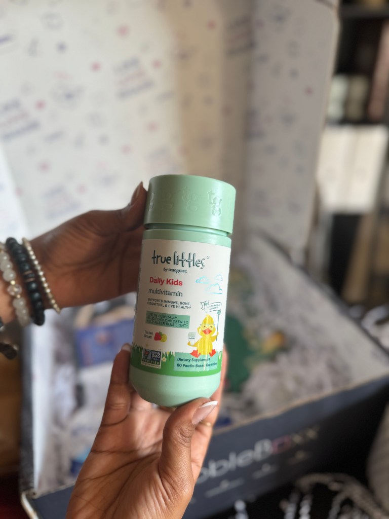A hand holds a bottle of True Littles Daily Kids Multivitamin, featuring branding elements like a cartoon character and colorful design, with a box in the background.