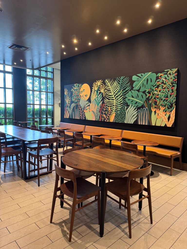 Interior of a café featuring a large mural of tropical plants on a dark wall, wooden tables and chairs arranged for dining, and large windows allowing natural light.