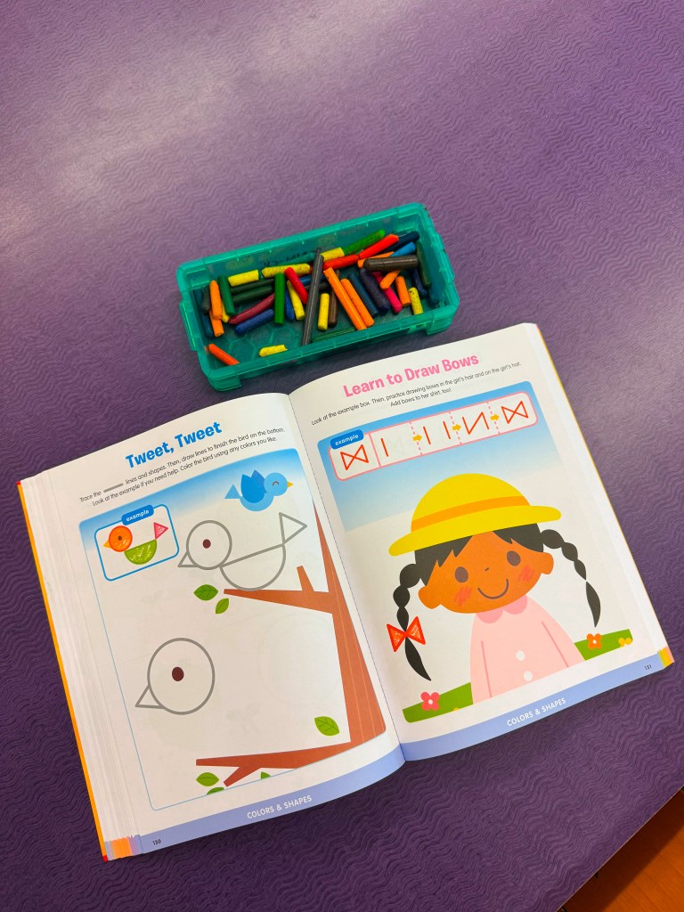 A children's activity book opened to pages about drawing, with illustrations of a bird on a branch and a girl in a yellow hat. A container of colorful crayons is placed above the book on a purple surface.
