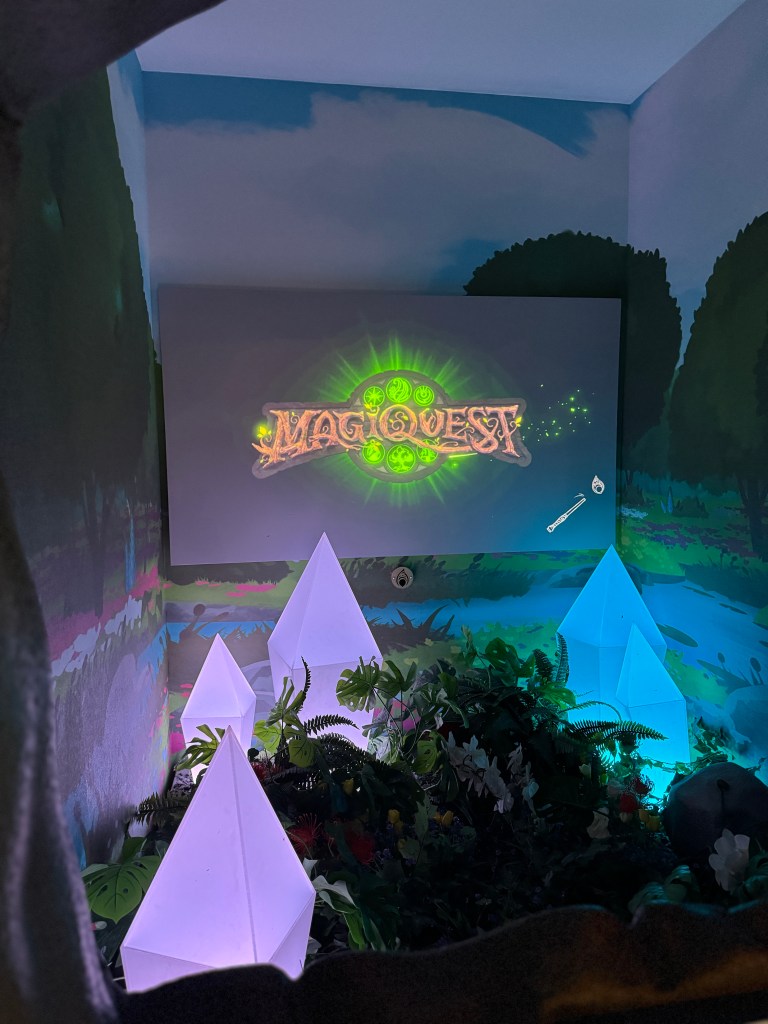 A colorful MagiQuest sign displayed in a lively themed area with ambient lighting and decorative elements including glowing triangular structures and foliage.