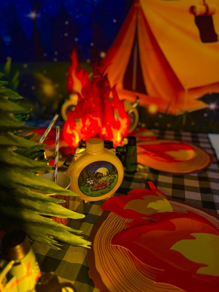 Nighttime camping birthday party table with glowing lanterns, campfire plates, pine trees, and tent backdrop.