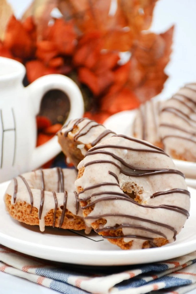 Close-up of baked pumpkin donuts topped with a creamy glaze and chocolate drizzle, served on a white plate with autumn leaves in the background.