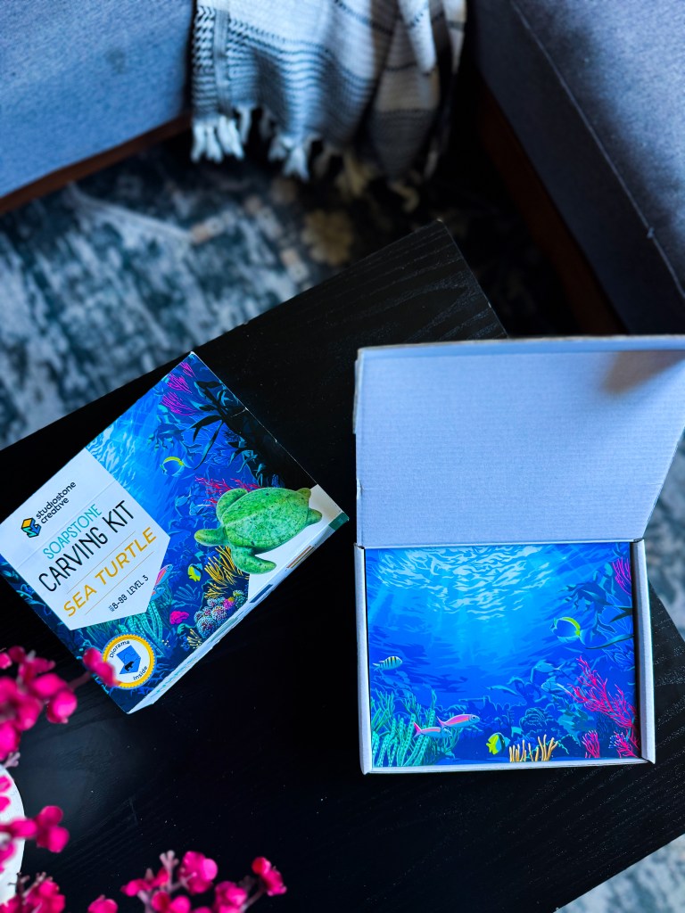A soapstone carving kit for a sea turtle, with an open box revealing colorful underwater scene artwork, placed on a black table with a pink flower arrangement.