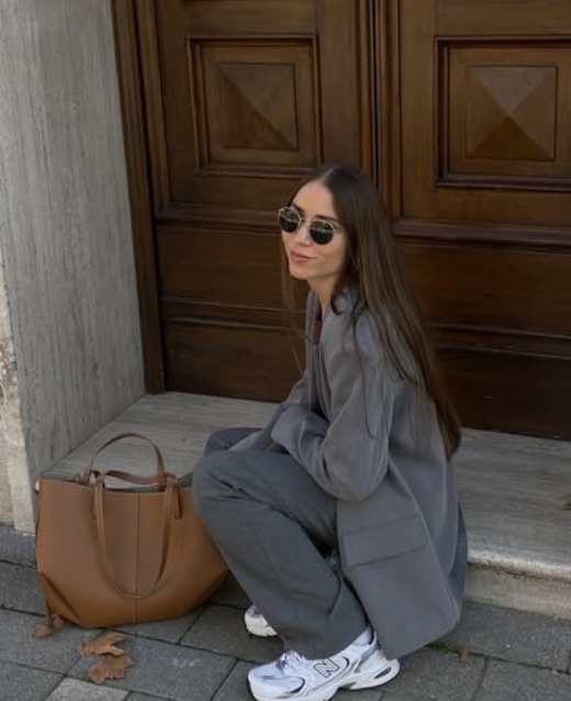 A woman squatting beside a brown handbag, wearing a gray oversized blazer, gray pants, and sneakers, sitting in front of a wooden door.