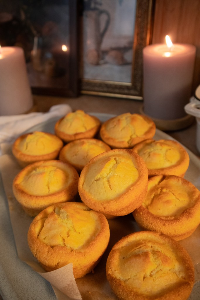 Freshly baked vegan cornbread muffins arranged on a plate, with candlelight and framed artwork in the background.