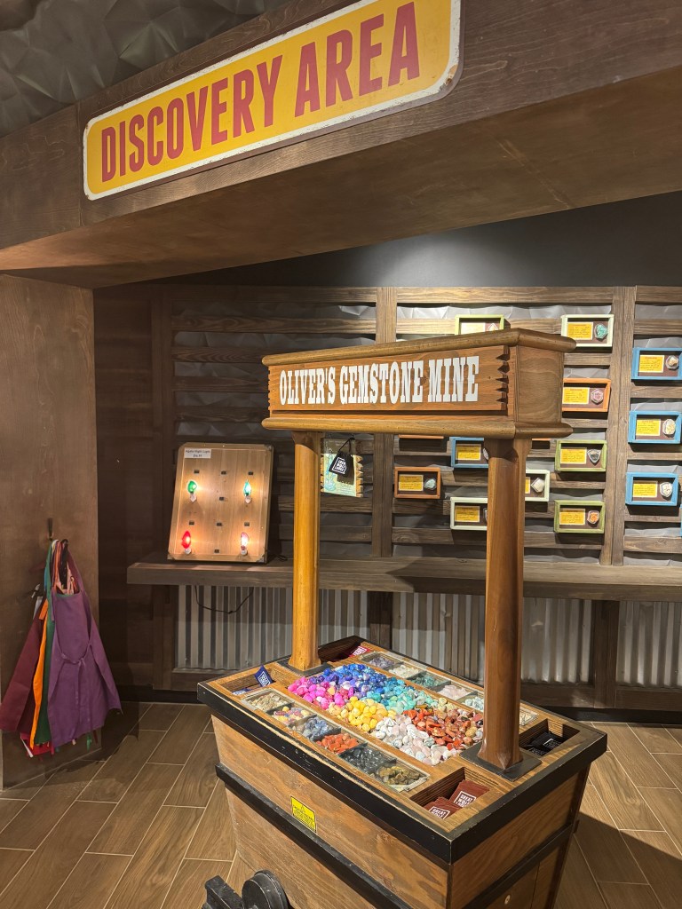 Interior view of the Discovery Area at Great Wolf Lodge, featuring a wooden cart labeled 'Oliver's Gemstone Mine' filled with colorful gemstones, with a backdrop of a wall displaying various gemstone information.