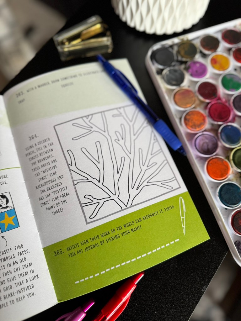 An open art journal page featuring a prompt to use colored pencils with an outlined drawing of branches, accompanied by watercolor paints, a pen, and a paper clip.