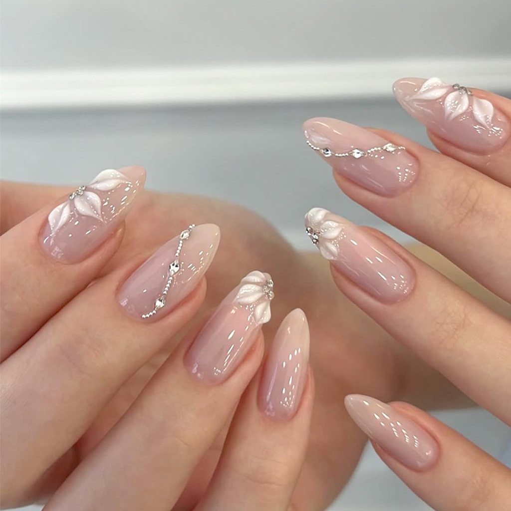 Close-up of elegantly manicured nails featuring a nude polish with white floral designs and silver embellishments.