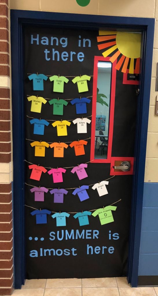 A decorated classroom door with colorful paper shirts hanging from string, displaying names and ages, against a black background. A sun cutout is near the top, with the text 'Hang in there... SUMMER is almost here' written in bright letters.