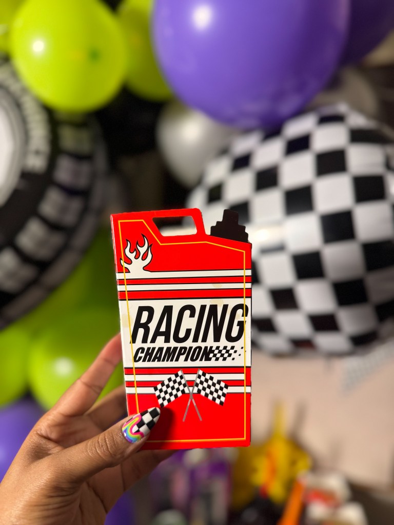 A person holding a red and black racing-themed treat case with the text 'RACING CHAMPION' and checkered flags in front of colorful race car birthday party balloons.