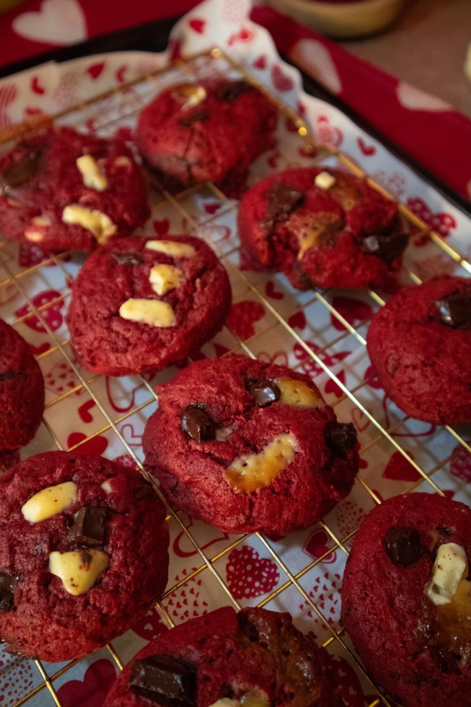 A close-up of freshly baked red velvet cookies on a gold wire rack, featuring chunks of white and dark chocolate, set against a decorative heart-patterned background.