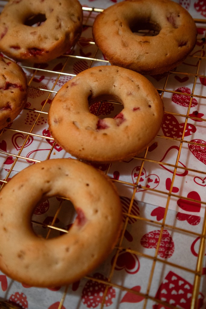 Freshly baked strawberry donuts cooling on a wire rack, with a decorative red heart-patterned background.