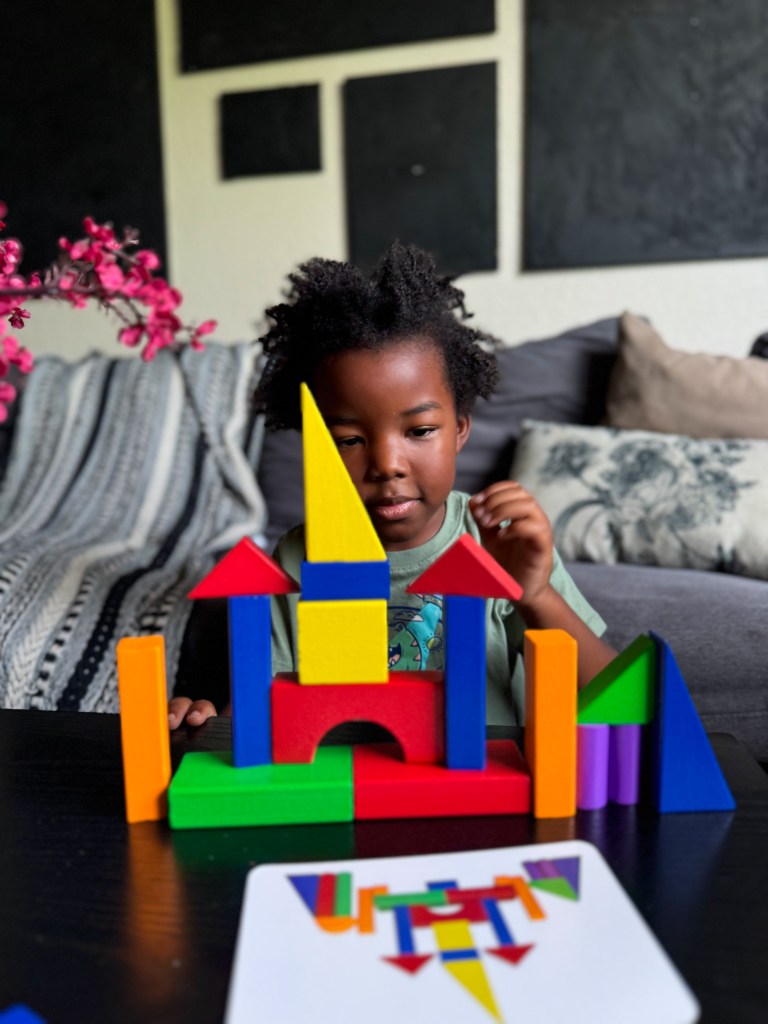 A young child building with colorful blocks, creating a castle-like structure, with a picture card of the design in front.