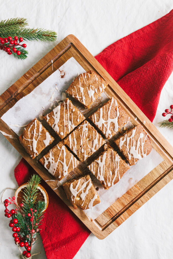 A wooden cutting board topped with nine squares of iced dessert, displayed on a white surface with a red cloth and holiday decorations.